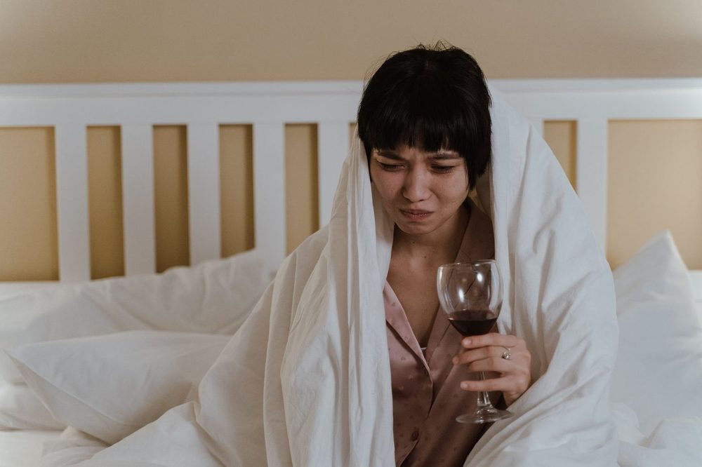 Zmęczona kobieta siedzi w łóżku otulona kołdrą, z kieliszkiem wina w dłoni – alkohol a sen