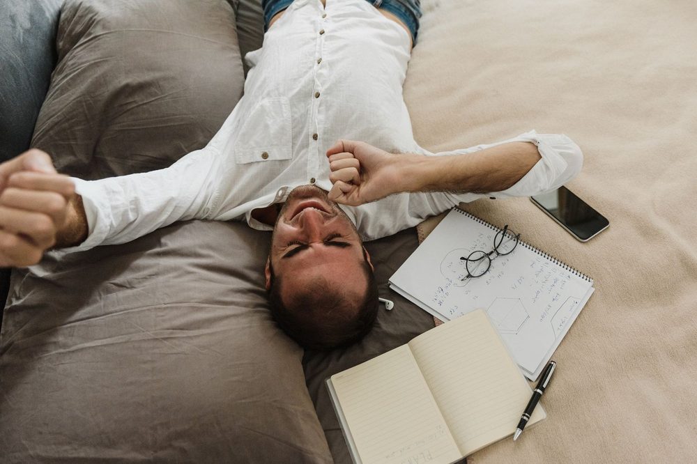 Homme fatigué allongé sur son lit parmi carnets et téléphone, le visage dans les mains — fatigue ou paresse