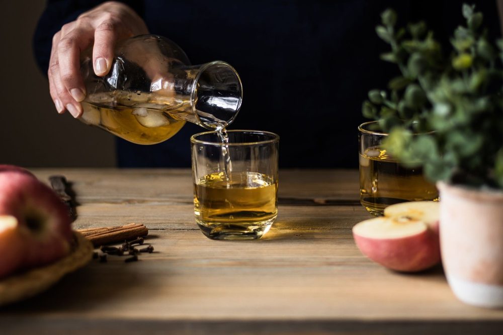 Goldene Flüssigkeit wird aus einer Karaffe in ein Glas neben aufgeschnittenen Äpfeln gegossen – Apfelessig trinken auf nüchternen Magen