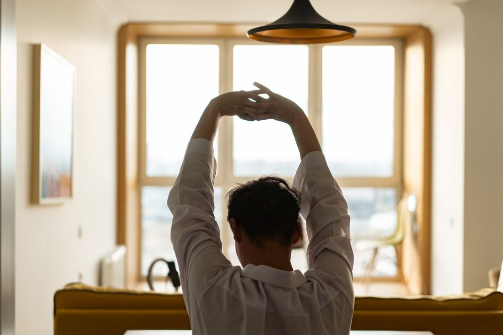 Mann streckt sich morgens am sonnendurchfluteten Fenster – Morgenroutine erfolgreicher Menschen und Energie am Morgen